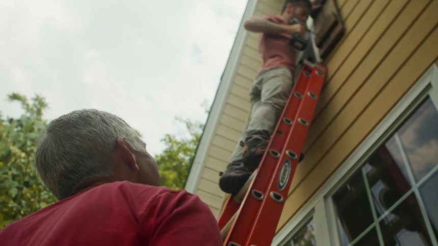 A carpenter drills into a wooden wall from a ladder outdoors while another person watches below