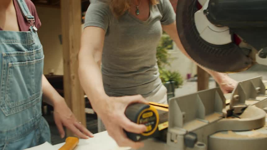 Three workers collaborate using a measuring tape and miter saw on a diy construction project
