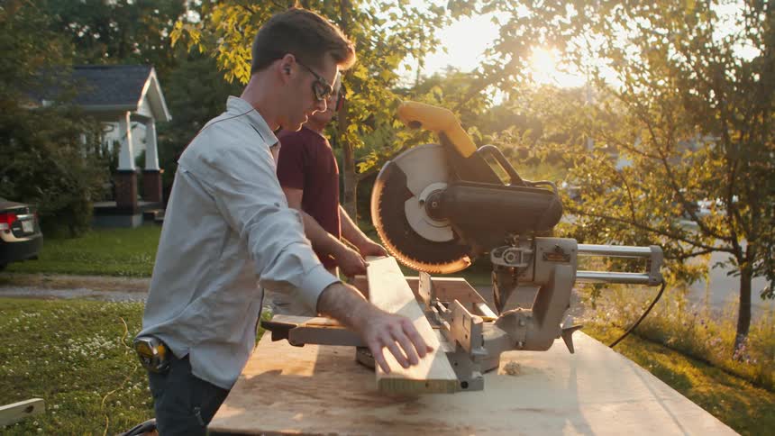 Two men carrying a piece of wood towards a circular saw