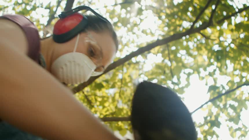 Woman in protective gear handling a circular saw on a wooden slat and other people in the background watching