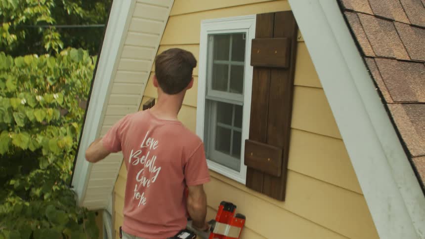 Man picking up a piece of varnished wood while standing on a ladder outside a house and placing it in a window