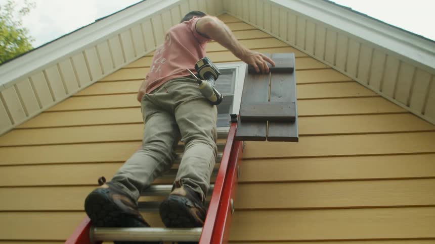 Man holding a window shutter climbing up a ladder outside towards a window