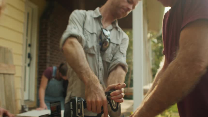 Man drills a screw into a wooden plank stabilised by two people on an outdoor worktop