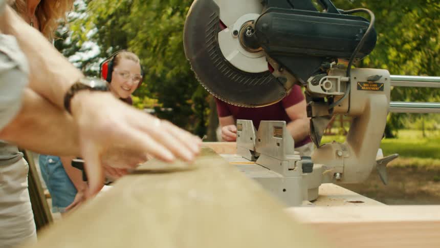 A man wearing safety glasses carries a wooden beam while three individuals use a miter saw to cut it outdoors