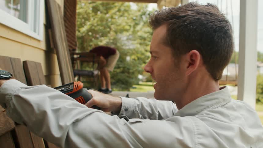 A man works in the front yard at midday driving a nail into a piece of wood with excitement nearby a dog on a leash wags its tail as the man continues his task