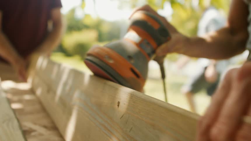 A person is using an electric sander on a wooden plank outdoors on a sunny day