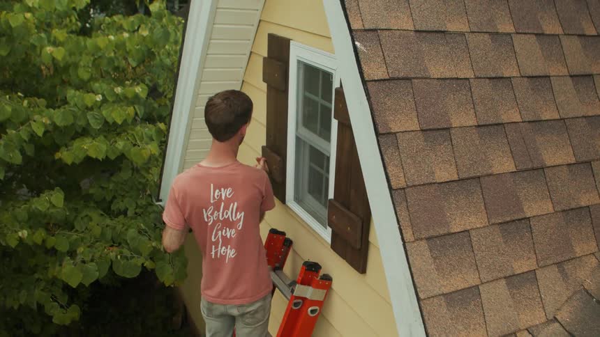 Carpenter drills while standing on a ladder as another person assists from below