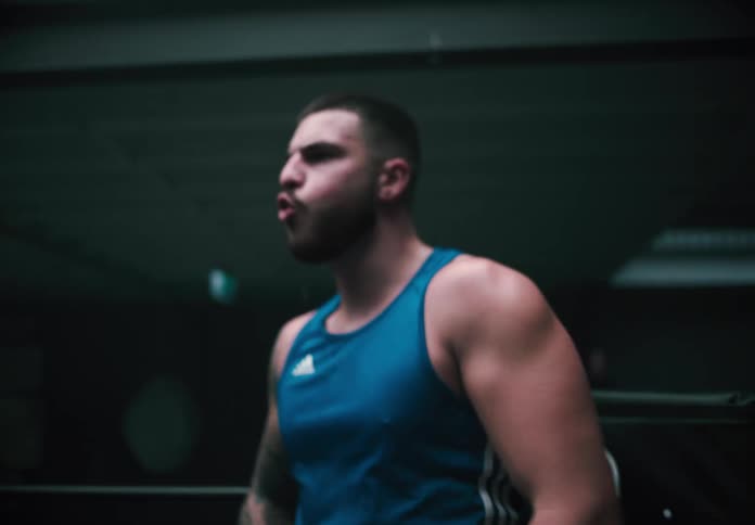 A boxer in a blue tank top warms up in the ring with his trainer standing behind him