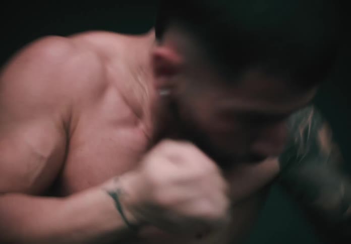 A shirtless man with a tattoo on his arm practices boxing under dim lights in a dark stadium