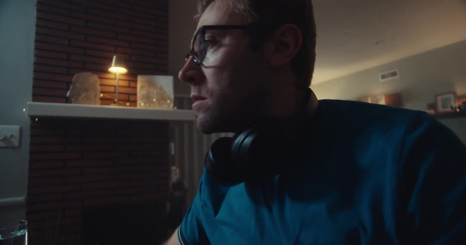 A man wearing headphones looks worried while sitting at his computer in a dimly lit room