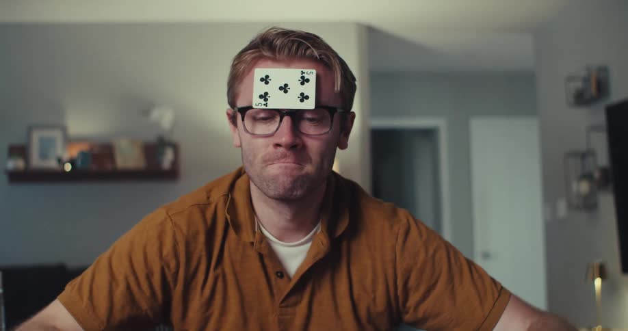 A man with a playing card stuck to his forehead appears frustrated while playing a card game indoors