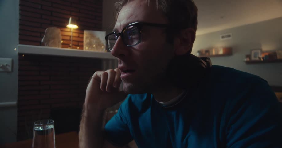 A fairskinned man with reddishblonde hair and glasses wearing headphones and a blue shirt sits at a desk with a computer his expression showing amused surprise