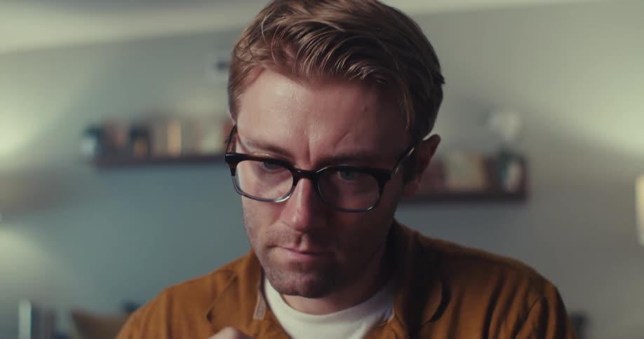 A fairskinned man with light brown hair and glasses sits indoors looking intently down at his cards