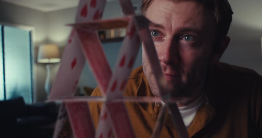 A fairskinned man with light brown hair and blue eyes looks at a precarious house of playing cards in a softly lit room
