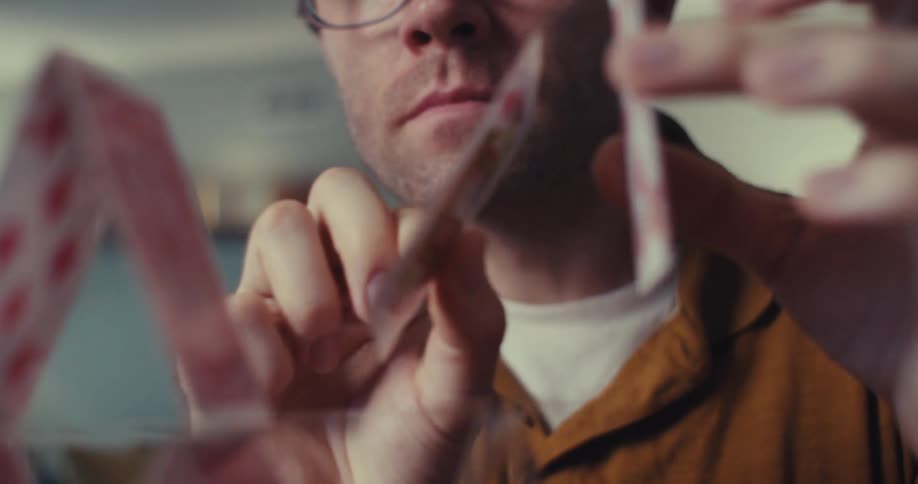 A fairskinned man with light brown hair carefully places a playing card onto a fragile house of cards