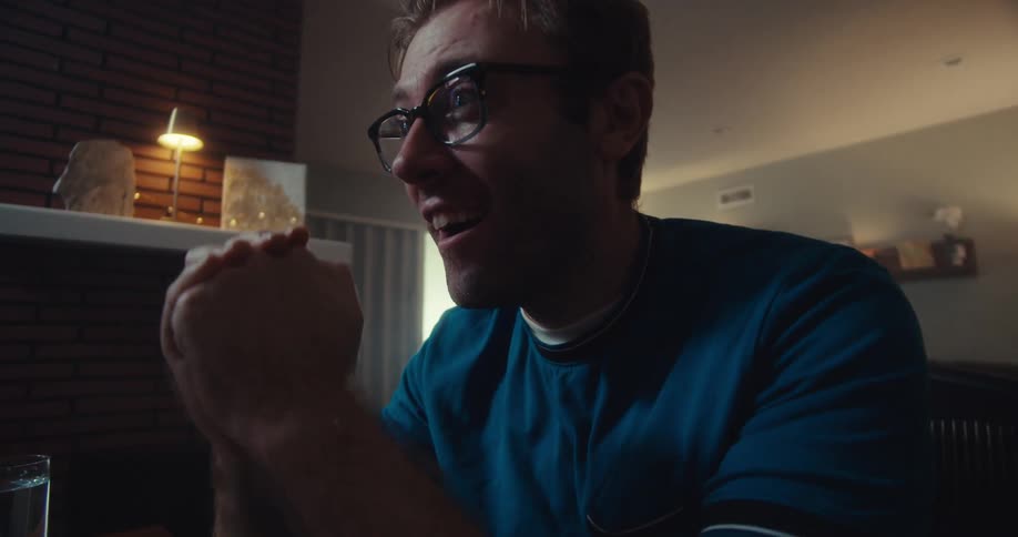 A man wearing glasses sitting in front of a screen with sudden excitement in his living room