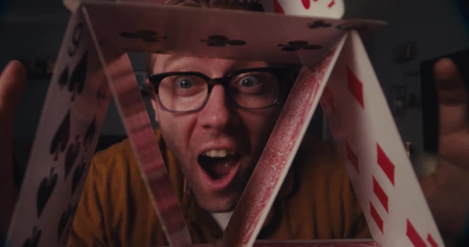 A proud man beams with joy as he peers through the pyramid of playing cards he just built indoors