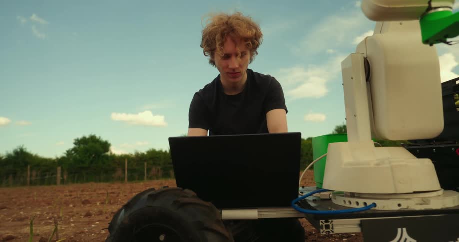 A man is using his laptop to work on a agriculture robot on the farm