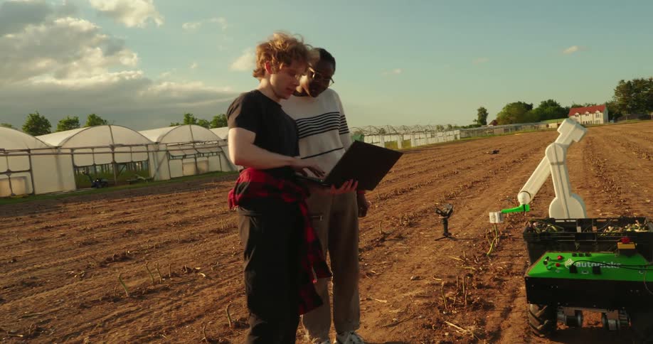 Two agriculture students researching in a field with a laptop by robotic machinery and greenhouses