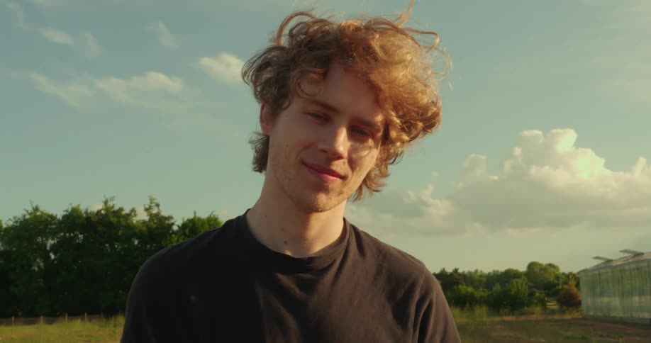A young man with curly blonde hair and blue eyes looks off to the side against a backdrop of a partly cloudy sky and trees