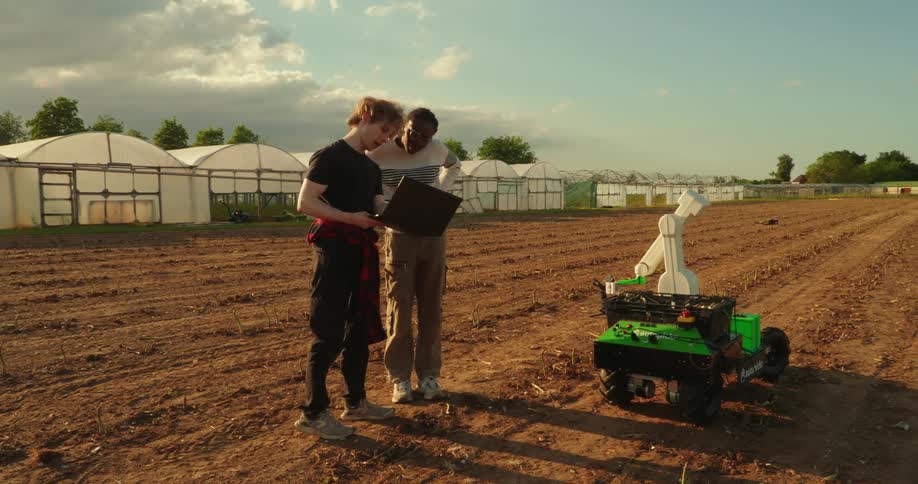 Agriculture students researching in a field with laptops and an agrirobot near a greenhouse