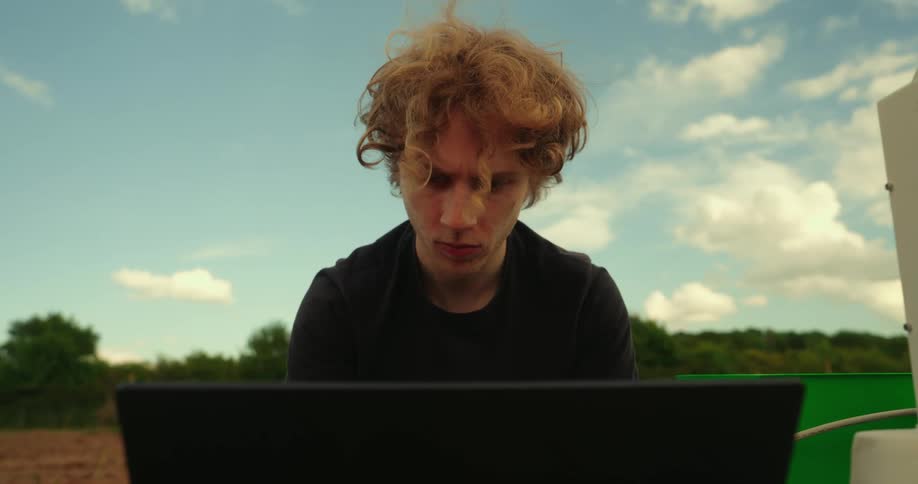 A young white man working on a computer in a field at midday