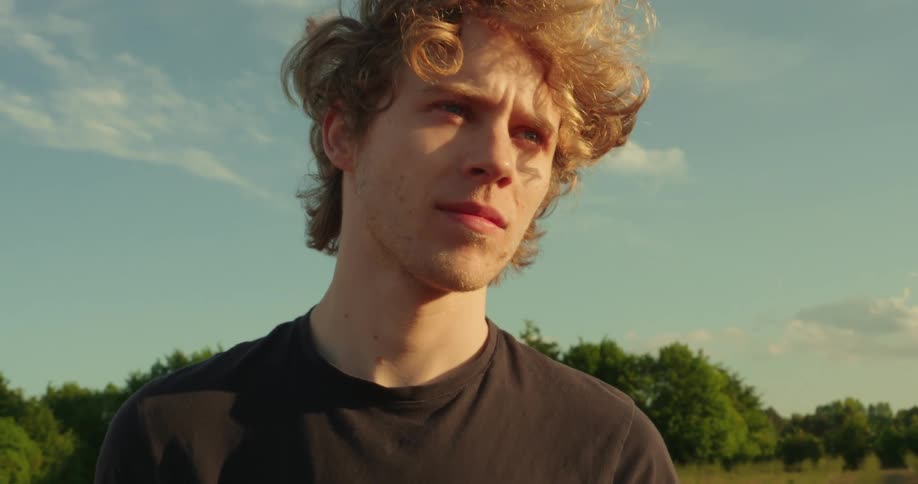 A young man with curly blonde hair and blue eyes looks off to the side against a backdrop of a partly cloudy sky and trees