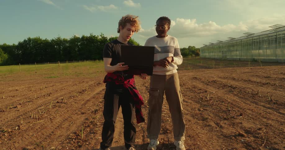 Two adults stand in a tilled field with a laptop discussing something under a partly cloudy sky near a large greenhouse