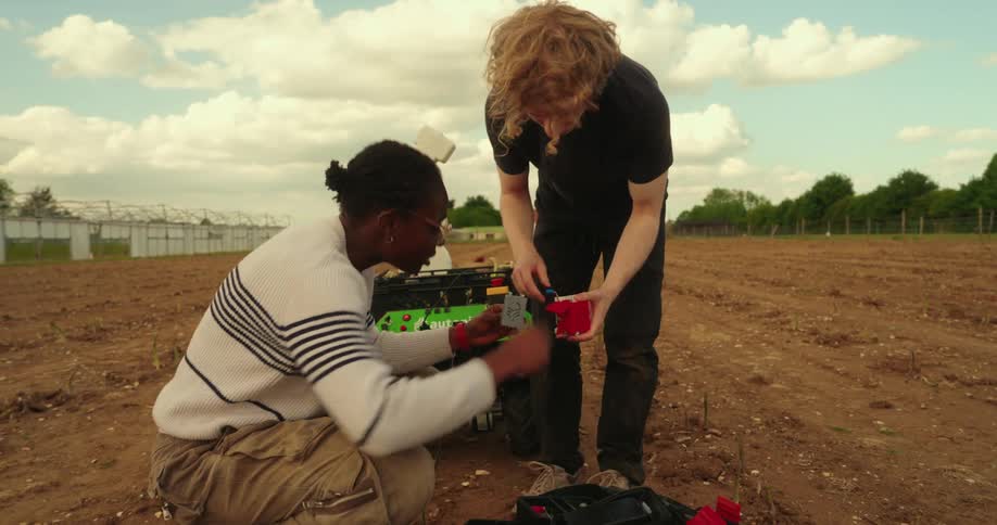 Two young adults are working on a small agricultural robot in a field with rows of plants under a partly cloudy sky