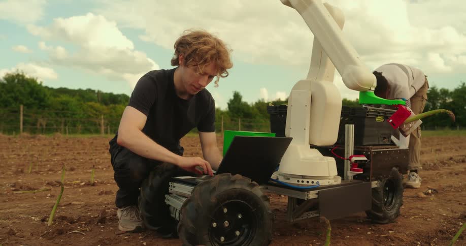 A man works on a laptop next to a piece of robotic farm equipment and another man walks up with a toolbox