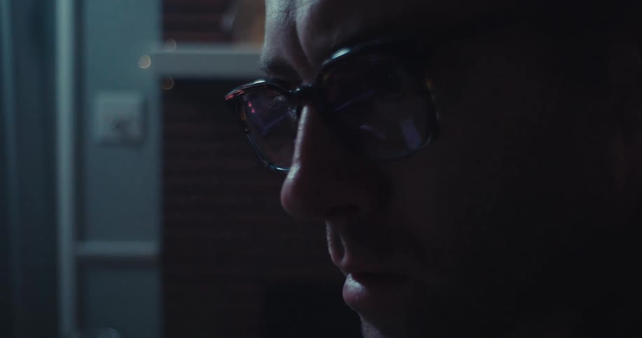 A man wearing glasses sits in front of a computer watching a stock market chart