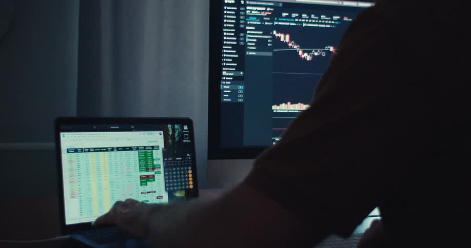 Man uses a laptop standing on a table next to a large computer monitor