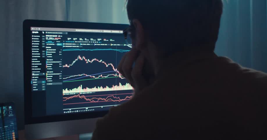 A man analyzes stock market charts on a computer screen in a dimly lit room