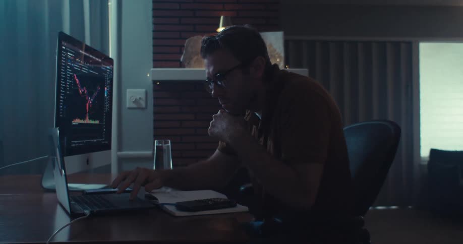 A man intensely analyzes stock data on both a computer and laptop in a dimly lit living room