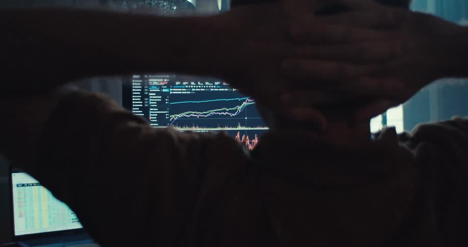 Close up of a man stretching and placing his hands behind his head while sitting in front of a trading desktop setup