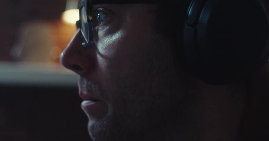 A man wearing reading glasses and headphones is making movements with his mouth and when he is looking forward