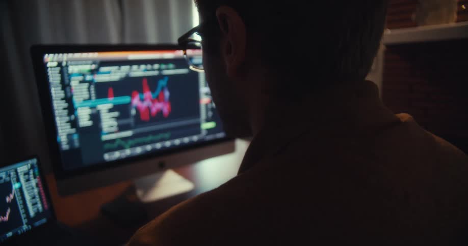 A man wearing glasses works on two computer screens displaying financial graphs in a dimly lit room