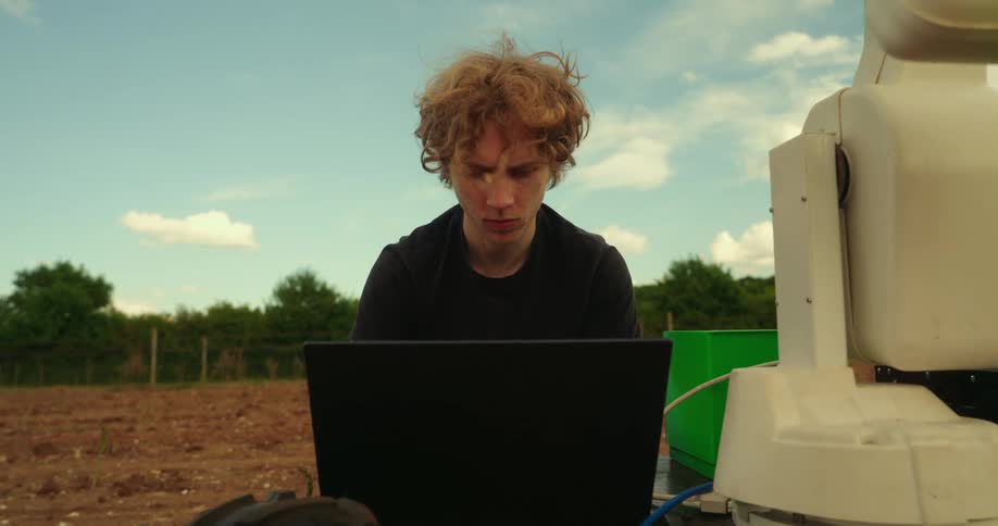 A man sits at a computer with his robot outside on a farm