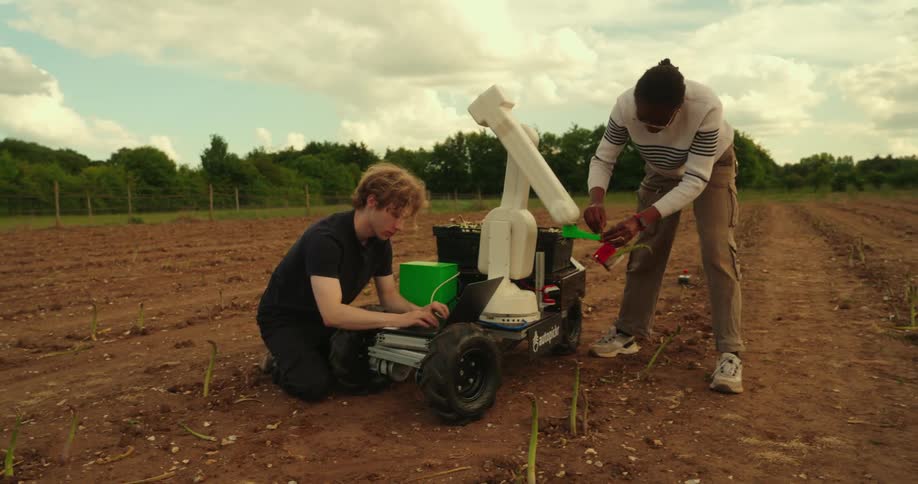 One person works on a laptop while another assists with a robotic farming device in a field