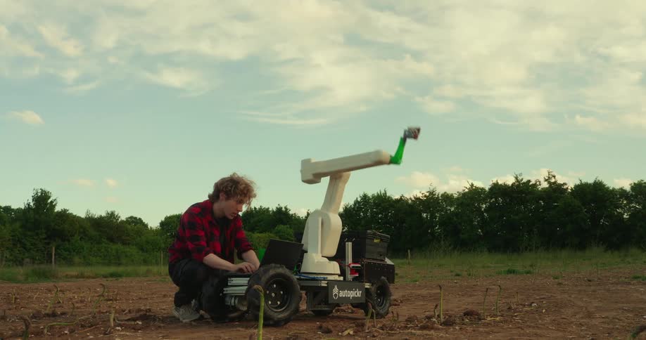 A young man in a plaid shirt kneels beside a small white robotic device with a moving arm operating it with a laptop in a field under a cloudy sky