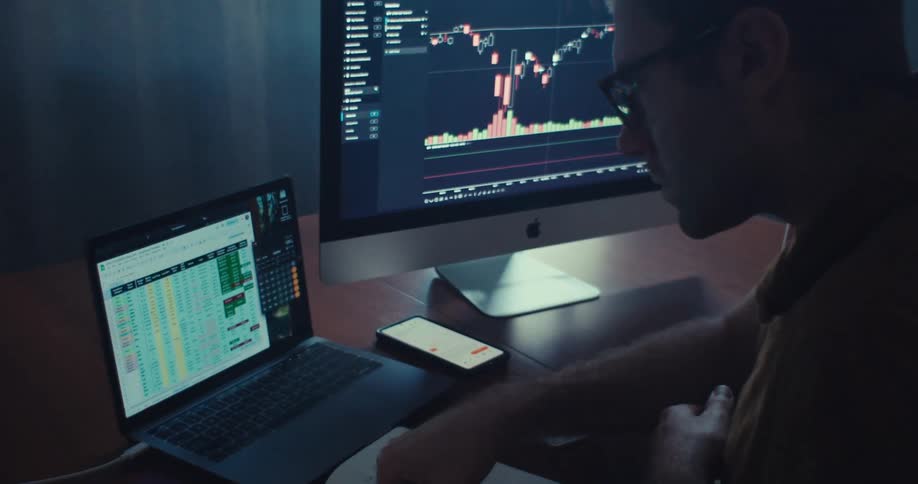 A man studies financial charts and spreadsheets on multiple screens in a dark office