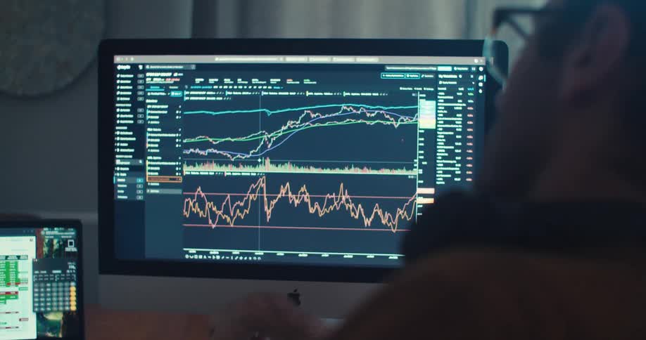 A man intently analyzes stock market charts on a computer screen in a home office setting