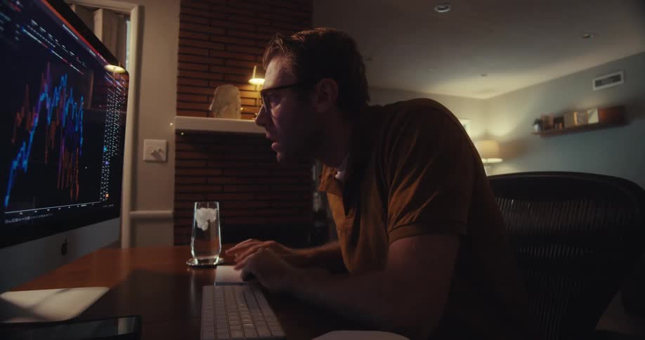 A man wearing glasses gets frustrated working on a computer displaying financial graphs at a desk in a dimly lit room at night