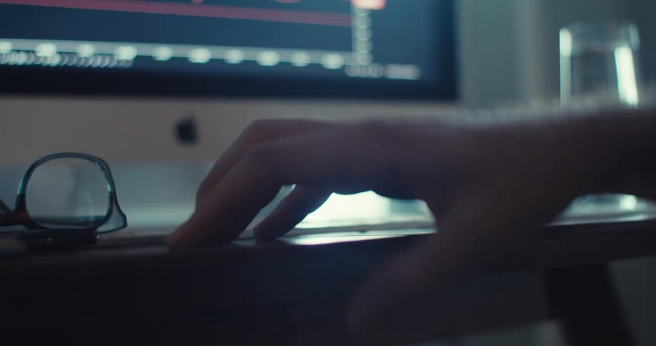 A man taps on a computer desk shifts his glasses to the side then starts typing on his keyboard while a stock market chart is displayed on the screen