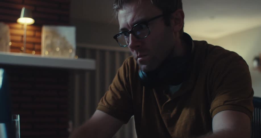Hands of a man with headphones around his neck type on a laptop keyboard in a dimly lit setting with a notebook and calculator nearby