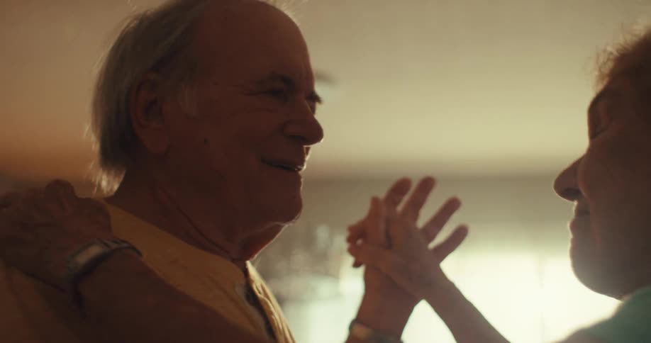 An elderly man dances with an elderly woman in the living room at midday
