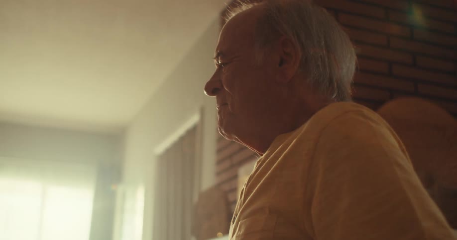 An elderly man in his living room shakes hands with another person as they slowly dance together at midday