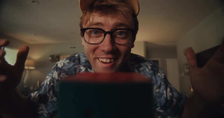 A man wearing glasses and a hat sits in his living room excitedly engaged with a game