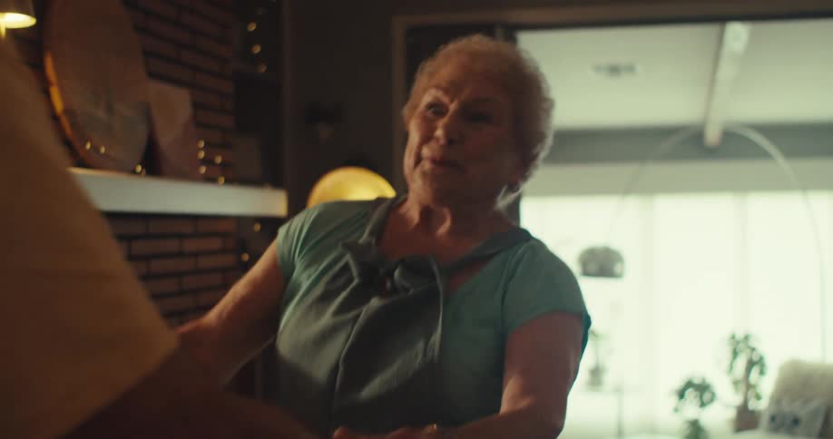 An older man laughs and looks off screen followed by an older woman in a teal top who smiles and talks engaging with him in a warmly lit room while dancing