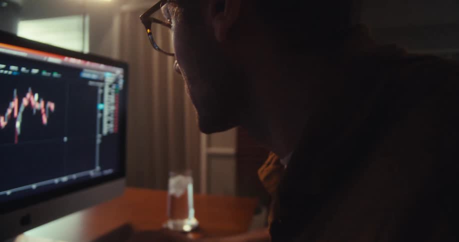 A fairskinned man with light brown hair and glasses looks at a computer screen displaying financial charts in a dimly lit room
